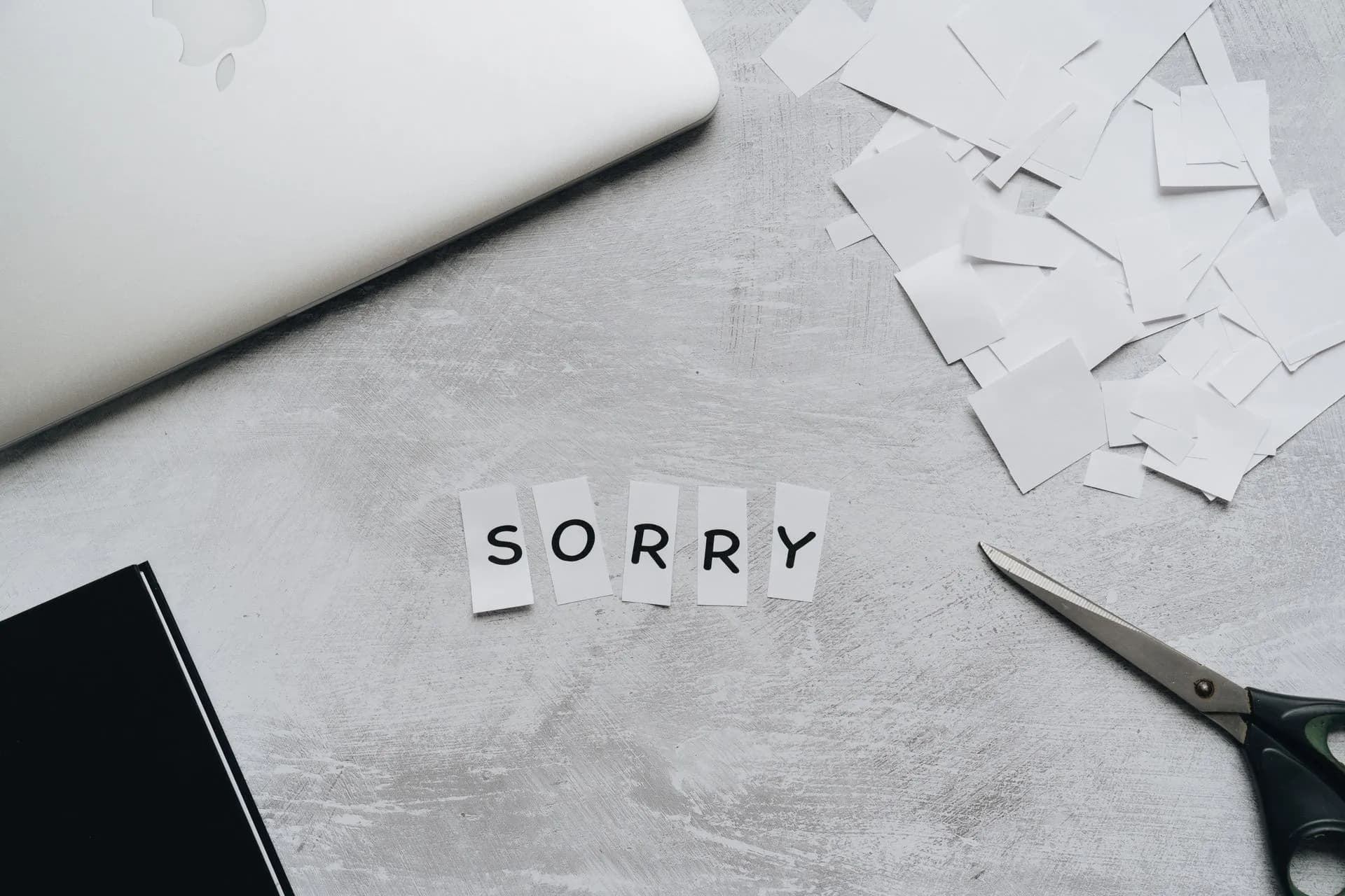 Top view of a desk with cut-out letters spelling 'sorry', scissors, and a laptop, illustrating California's SB 947 limits AI firings as NYC weighs AI safety bills and insurers eye CEO