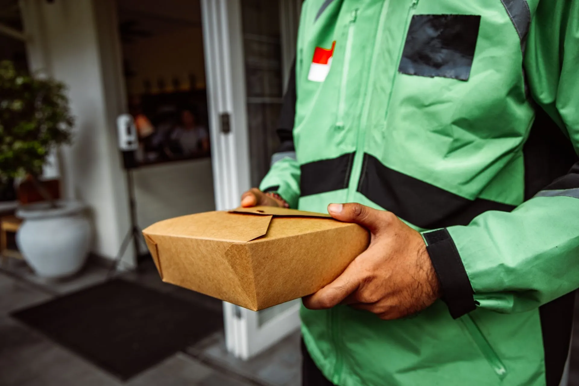 A deliveryman in a green jacket holding an eco-friendly food package outside a restaurant.