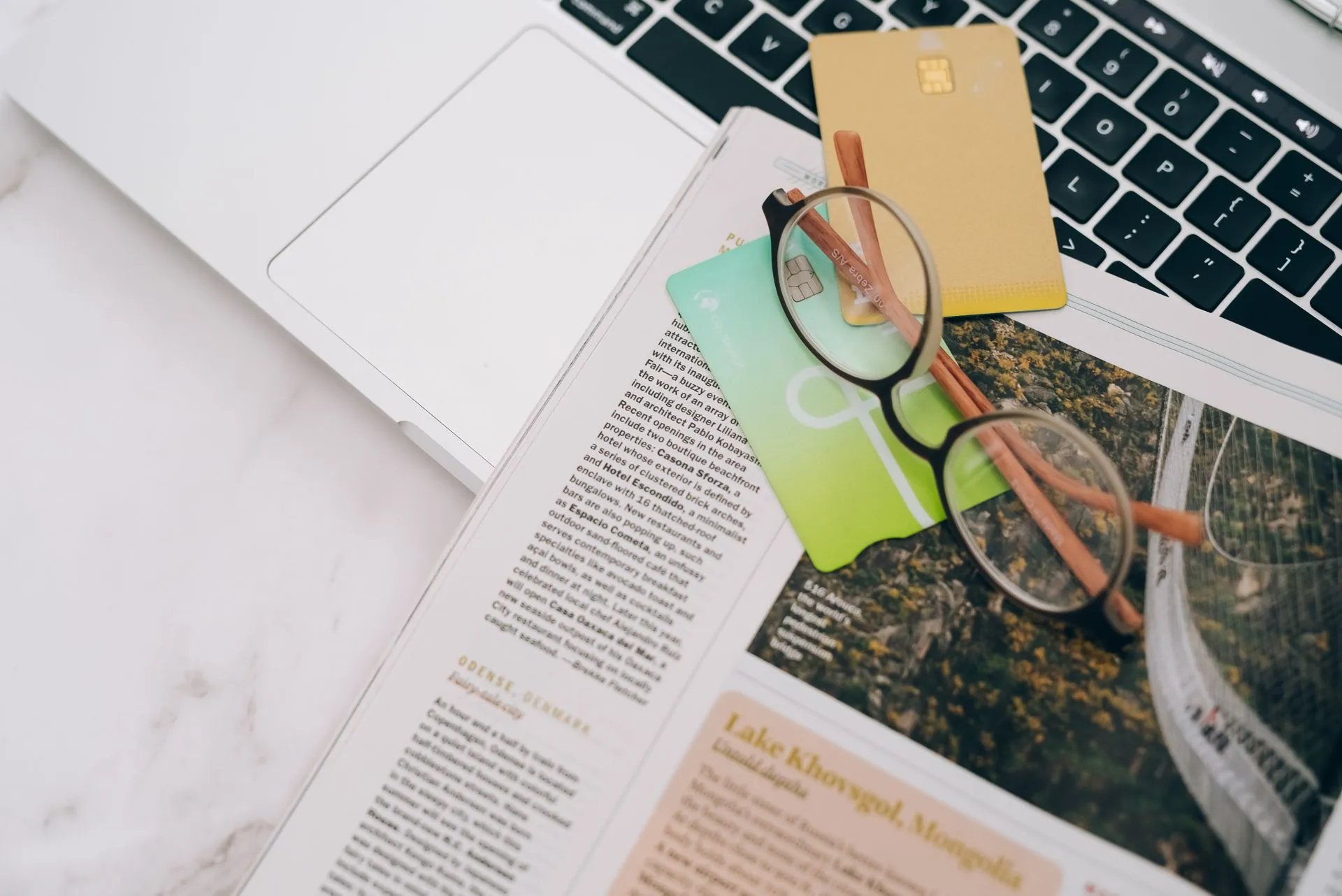 Eyeglasses resting on a magazine beside a laptop and credit cards, indoors, illustrating American Express resolution plan doubted by Fed and FDIC as Comptroller Gould votes no