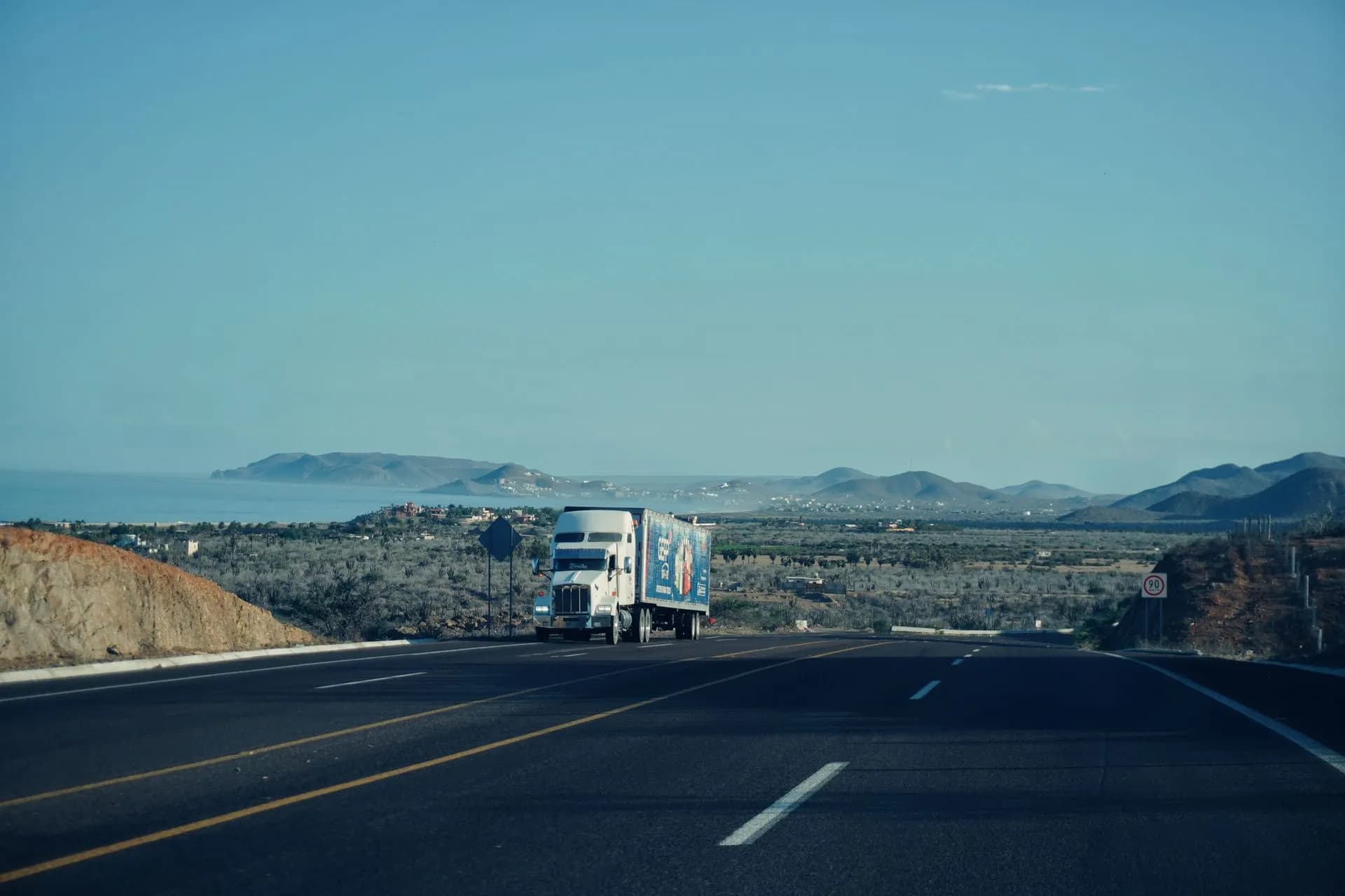 A semi-truck travels on a scenic highway with mountains in the background under a clear sky, illustrating Aurora driverless trucks