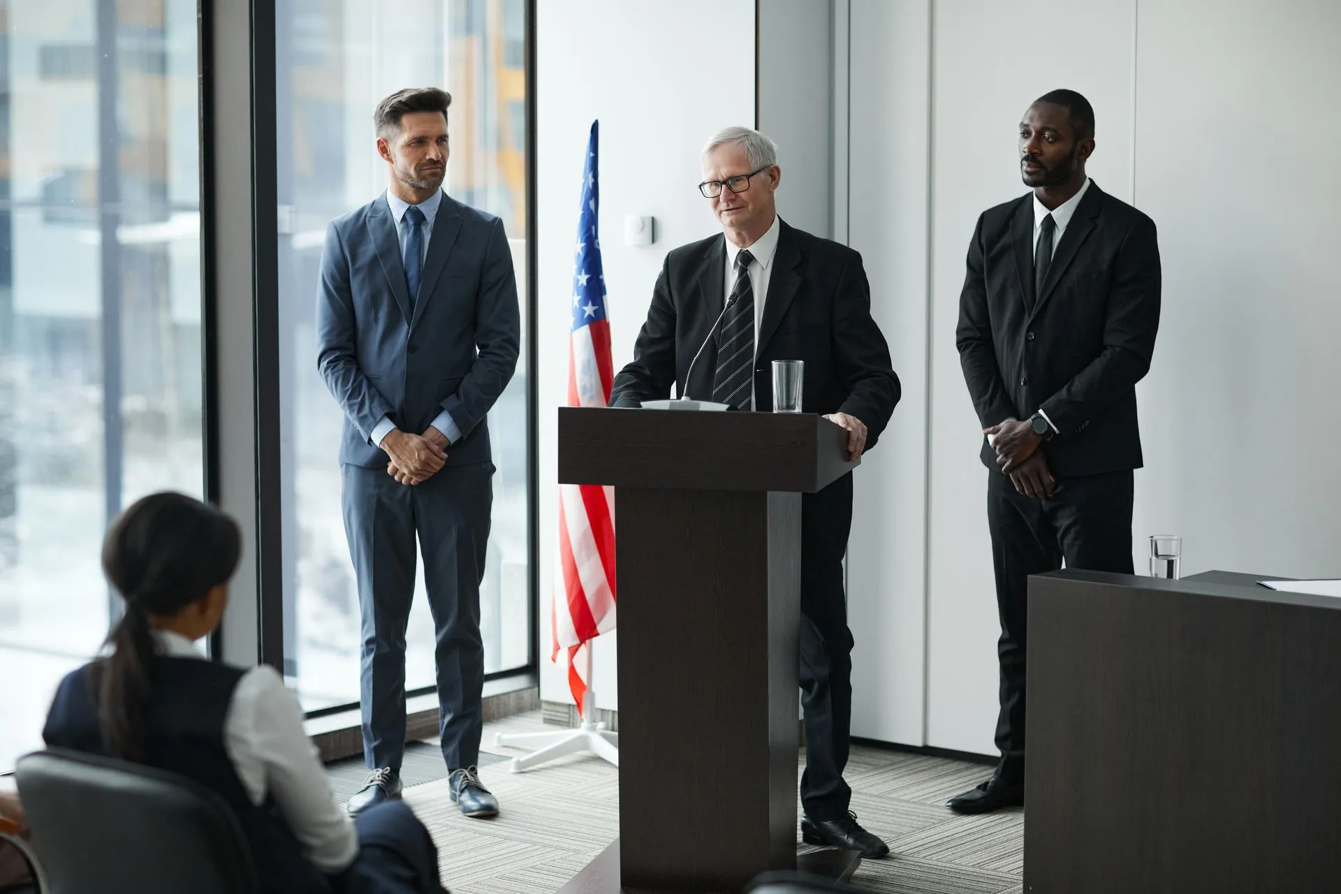 Officials delivering a political speech in a modern conference room with an American flag, illustrating CFTC staff say mention markets may be presumed susceptible to manipulation