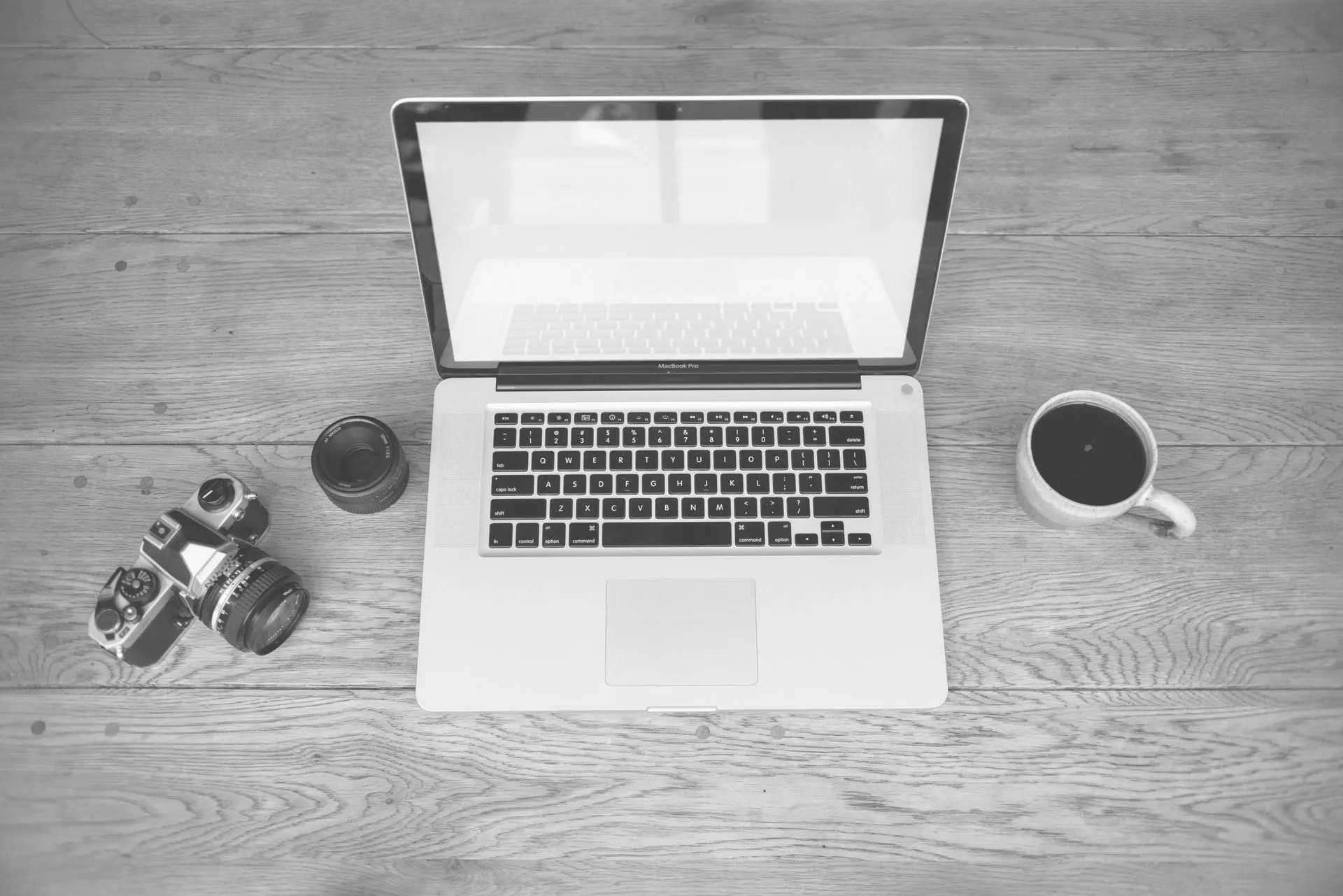 Monochrome image of a laptop, camera, lens, and coffee cup on a wooden desk, illustrating Cloudflare OS opens waitlist for a managed AI agent workspace for companies