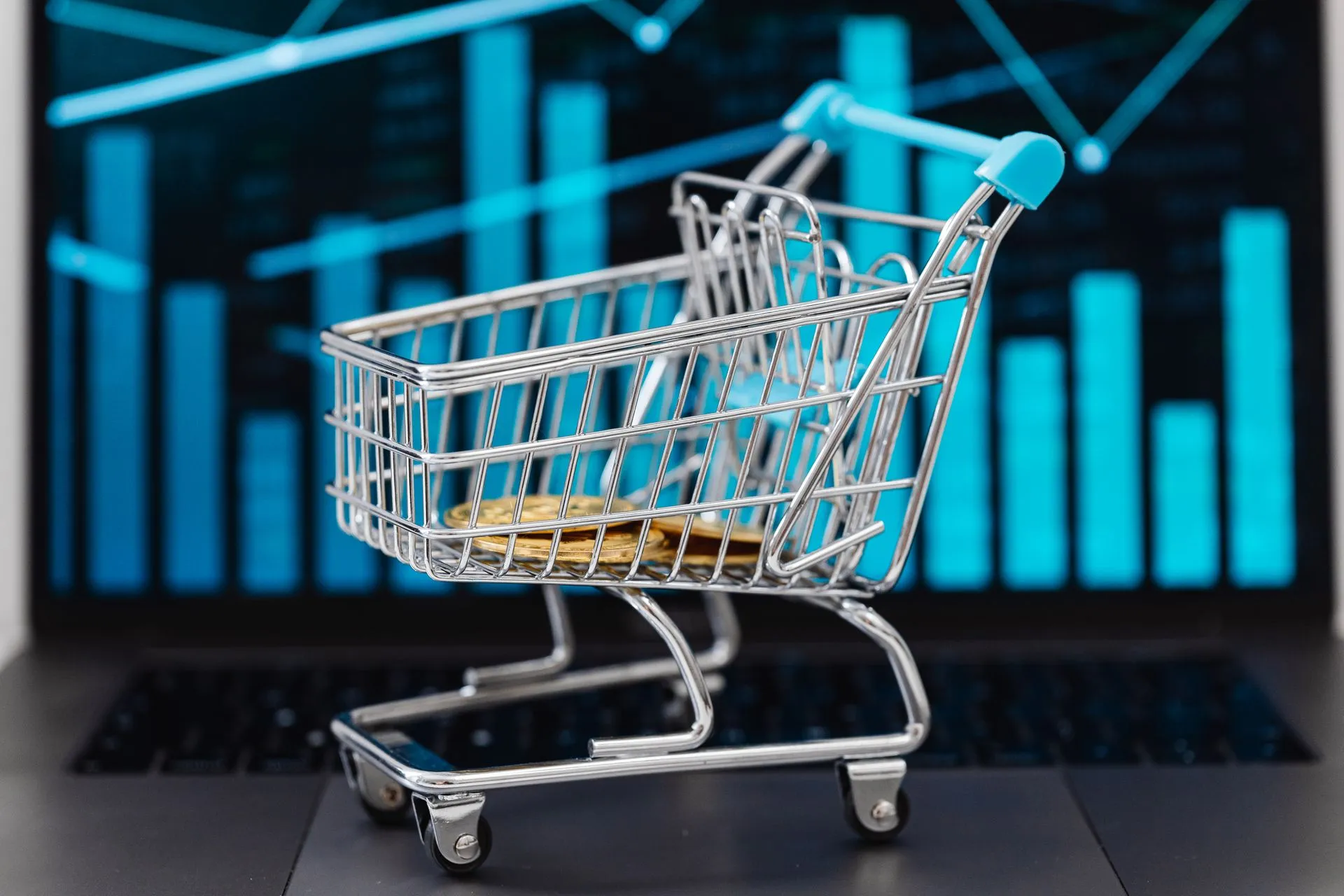A miniature shopping cart with coins sits on a laptop displaying financial graphs, symbolizing e-commerce and online shopping, illustrating Commerce launches catalog enrichment for BigCommerce and
