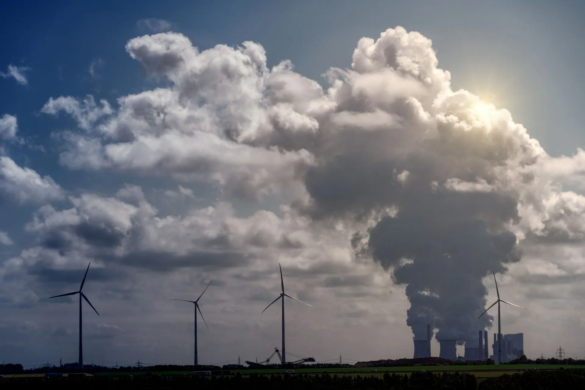 Wind turbines and a power plant under dramatic cumulus clouds in a rural landscape.