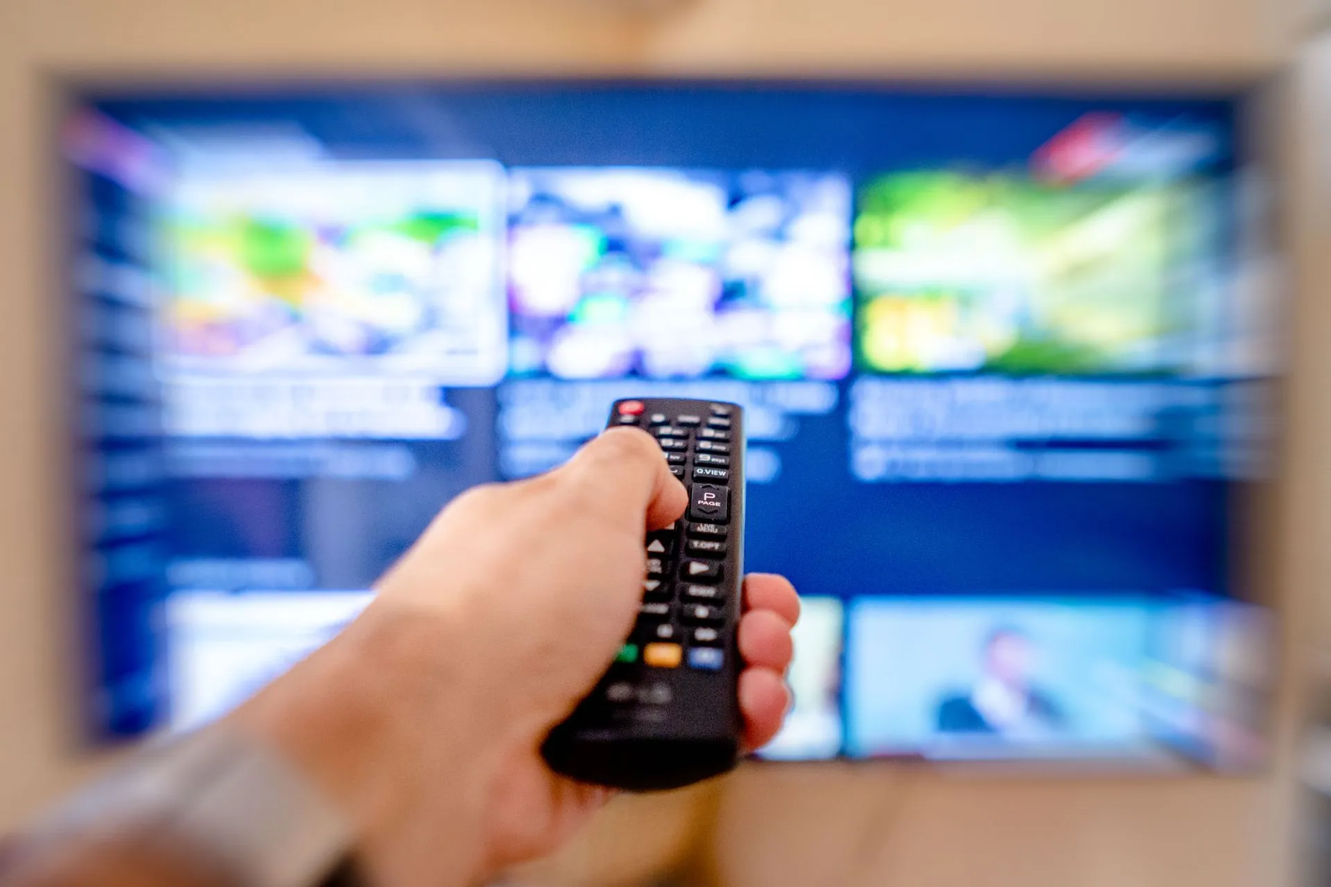 Hand holding remote control pointing at a blurred TV screen in a cozy indoor setting, illustrating Disney licenses Percy Jackson, Ice Age and Pixar films to Netflix in non-exclusive