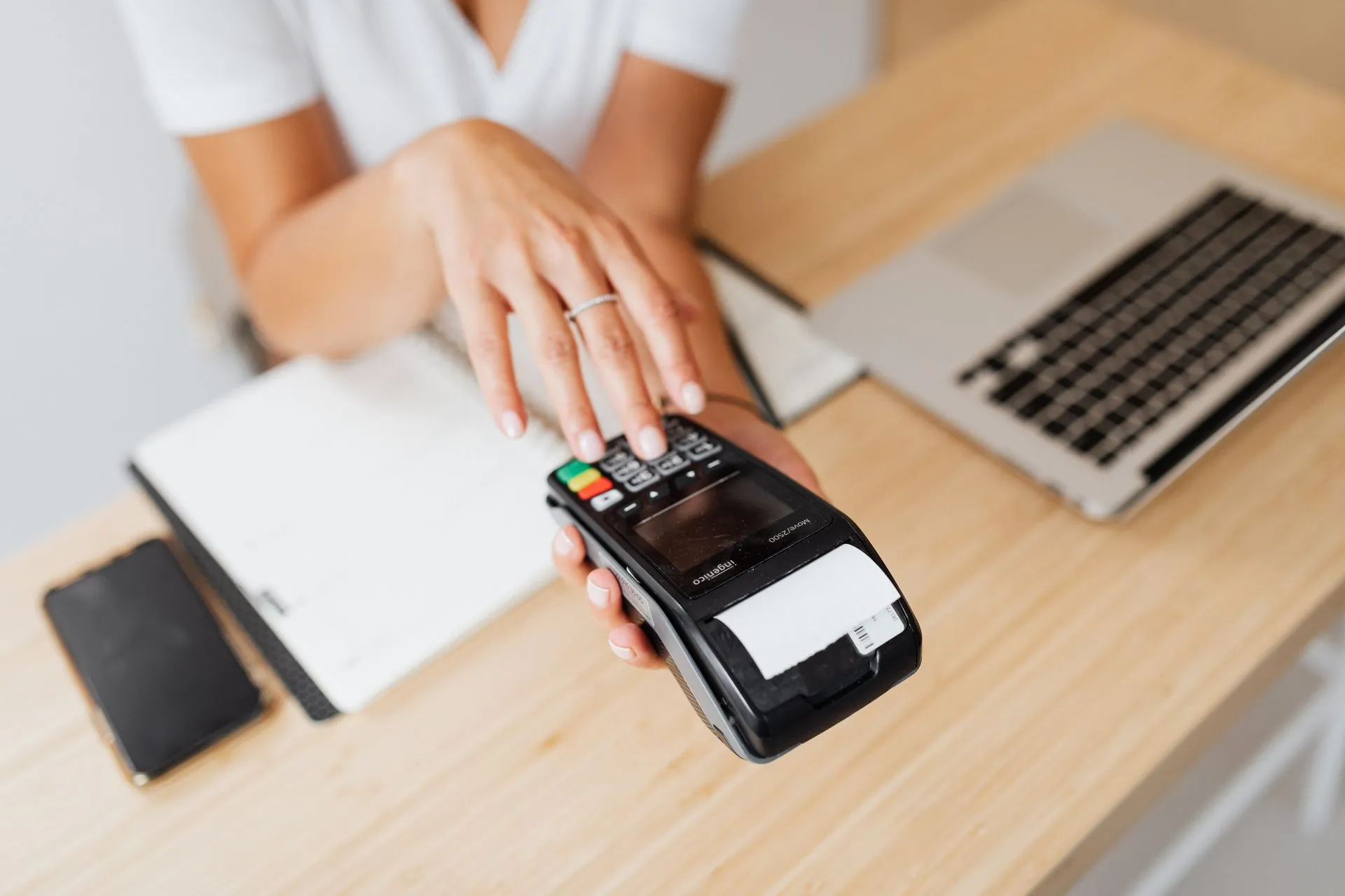 Close-up of a hand using a payment terminal beside a laptop on a wooden countertop, illustrating Wero, Bizum and Bancomat link up to reach 130 million users and challenge Visa and