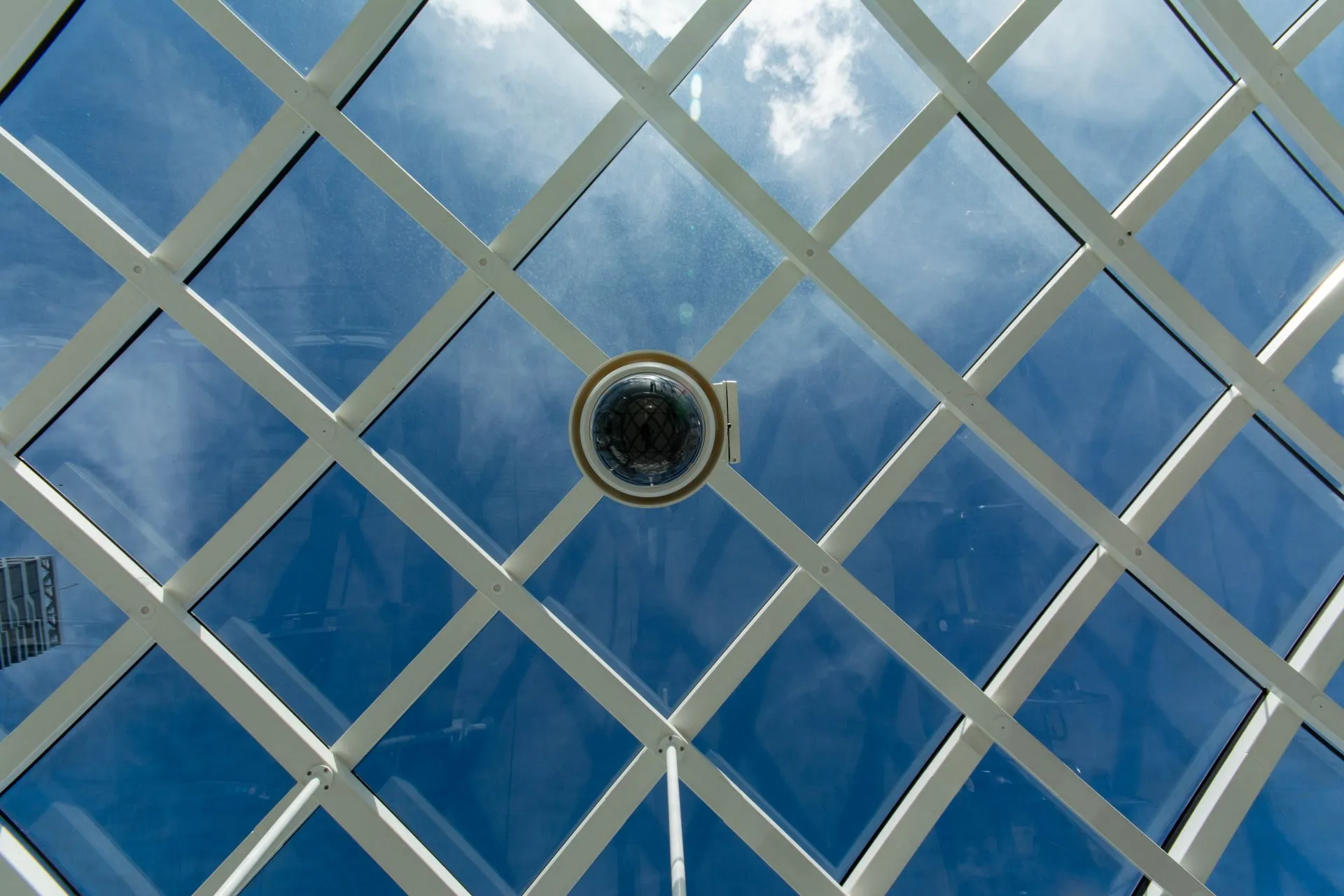 Surveillance camera mounted on a modern glass ceiling with sky reflections, illustrating Meadow AI raises $7 million for a digital secret shopper that watches and listens to staff