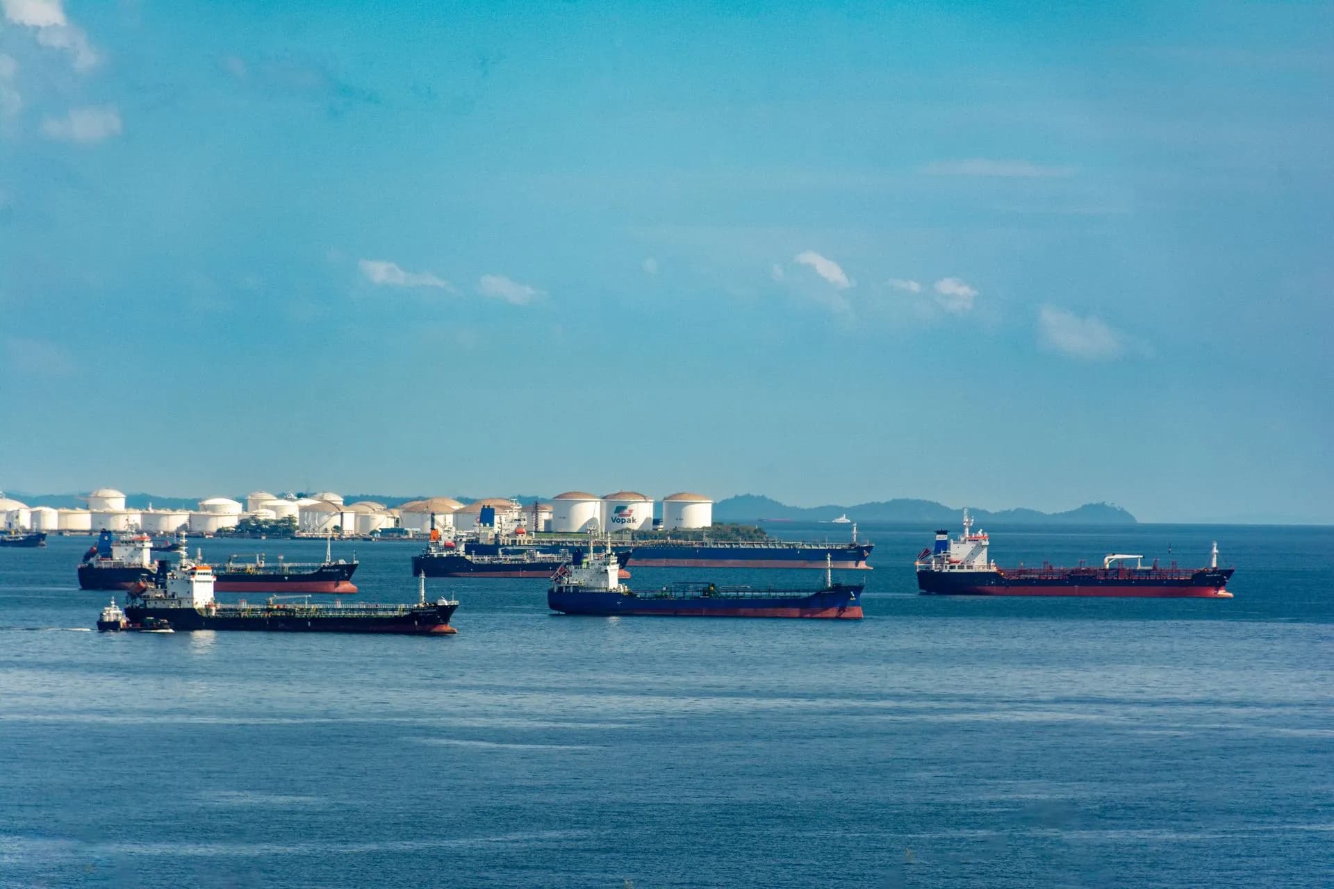 A fleet of cargo ships docked near oil storage tanks along a serene coastline with a clear blue sky above, illustrating Tanker cyberattacks prompt Coast Guard and FBI to board two