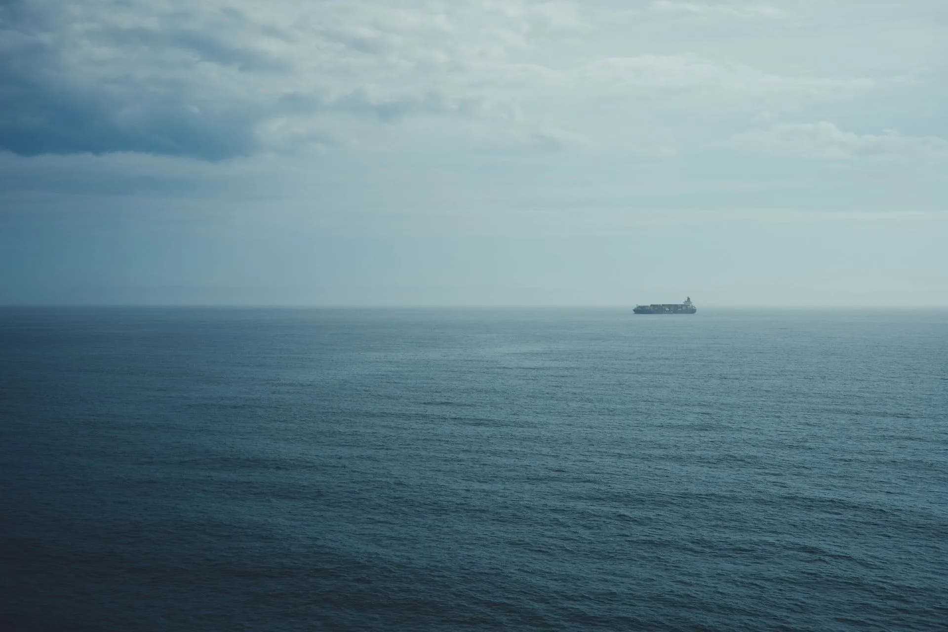 A distant cargo ship navigates through the calm, expansive ocean beneath a cloudy sky, evoking solitude, illustrating Quartermaster raises $140 million to put sensors on ships and map