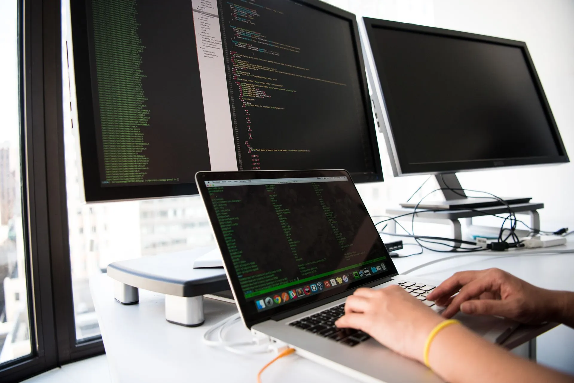 A developer writes code on a laptop in front of multiple monitors in an office setting, illustrating SonarQube Server 2026.5 LTA brings Sonar's AI code agents to companies' own