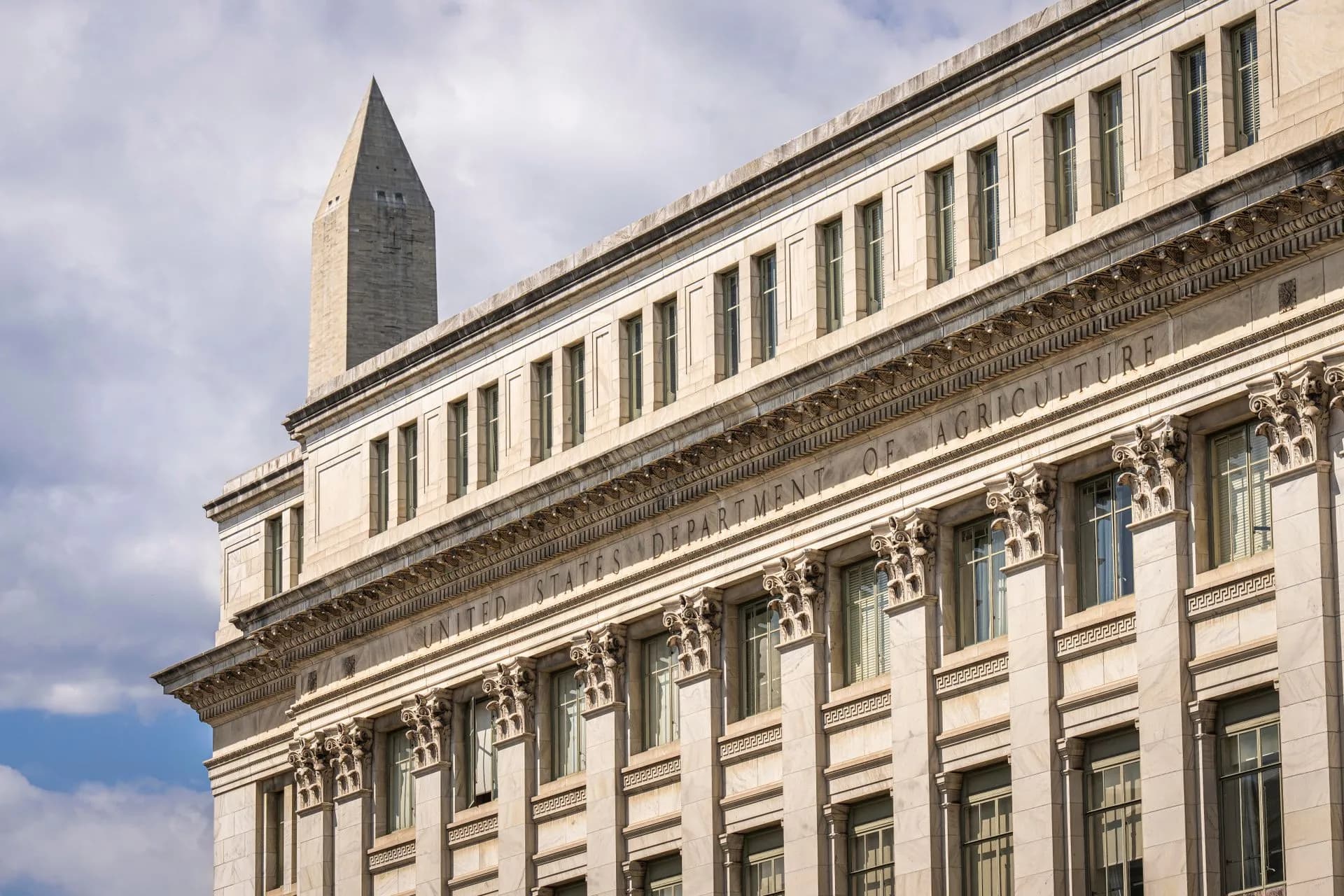 US Department of Agriculture building with Washington Monument behind, blue sky with clouds, illustrating Trump's Super Intelligence Force puts DNI Jay Clayton in charge of a 120-day