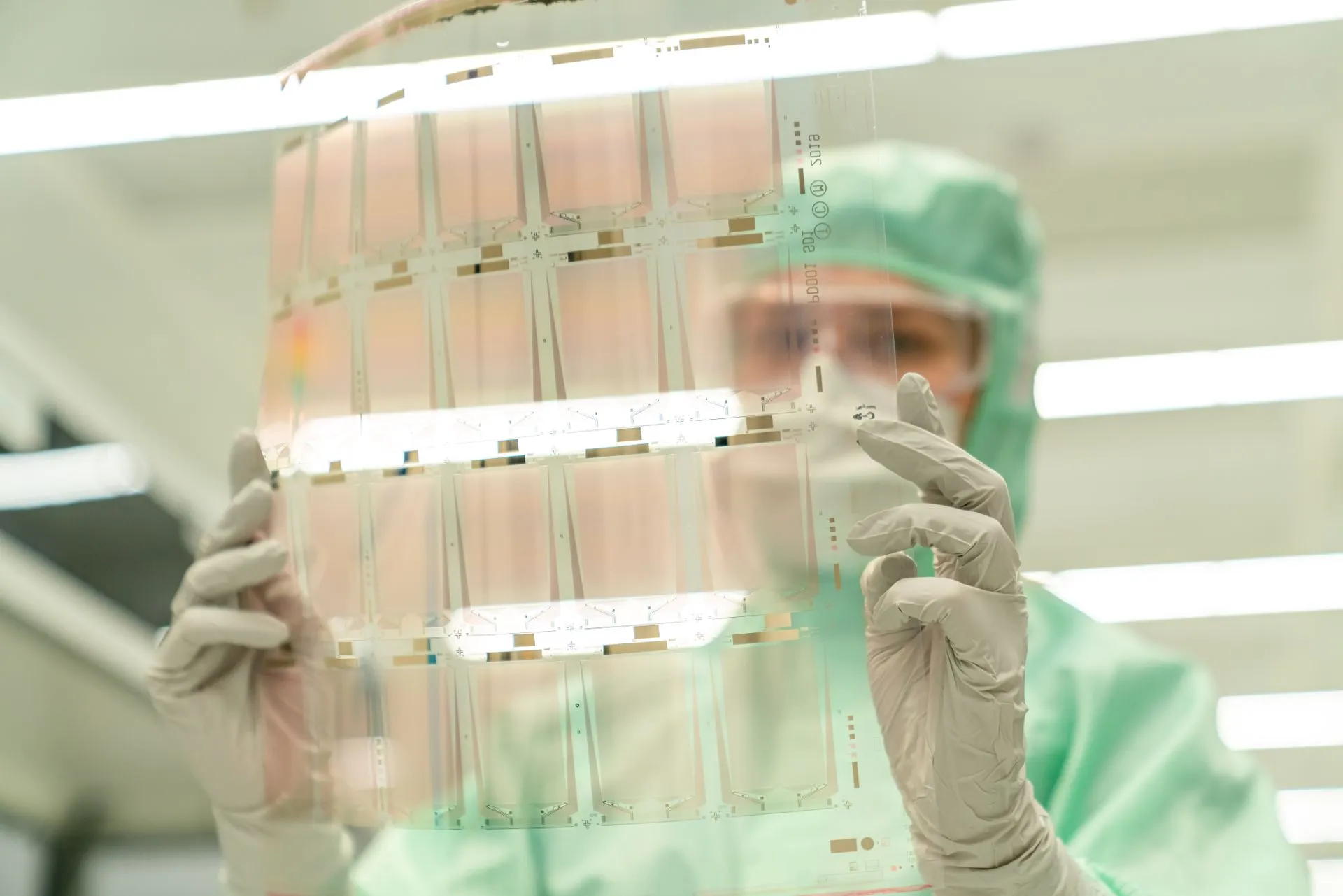 Scientist in protective gear holding a transparent test sheet in a laboratory, illustrating TSMC taps GlobalFoundries for US silicon interposers in a $2 billion, five-year deal
